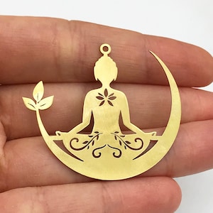Peut inclure: Pendentif en métal doré avec un motif découpé d'une personne assise en position de lotus sur une lune croissante. La personne a un motif de fleur sur le dos et il y a des feuilles et des volutes autour d'elle.