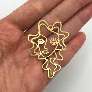 May include: Gold-toned metal cutout of a stylized face with wavy hair. The face has a single eye, a nose, and a mouth.