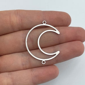 Peut inclure: Un pendentif en forme de croissant de lune en métal argenté avec un design à cadre ouvert et fin. La pièce comporte deux petites boucles circulaires, une située en haut et une en bas, permettant de l'attacher à des bijoux ou autres créations.
