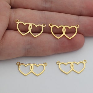 May include: Four gold-colored metal charms in the shape of two interlocking hearts. The charms are shown on a hand.