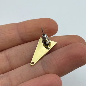 May include: A single gold-colored triangle earring stud with a silver post and backing. The earring is held in a hand against a white background. The earring has a small hole at the bottom corner.