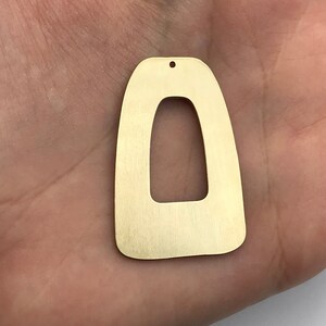 Könnte beinhalten: Ein goldfarbener Metallohrring mit einer einzigartigen, geometrischen Form. Der Ohrring hat eine große, offene ovale Form mit einer kleineren, rechteckigen Öffnung in der Mitte. Der Ohrring ist gebürstet für ein mattes Finish.