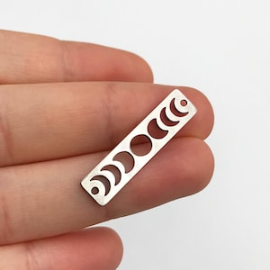 May include: A silver-colored rectangular charm featuring a series of crescent moon phases cut out along its length. The charm has a small hole at one end for attachment. The design is minimalist and celestial.