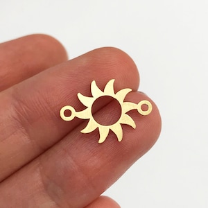 May include: A small, flat, gold-coloured metal sun charm featuring a central circular cutout surrounded by radiating curved rays. The sun design includes two small circular loops on opposite sides, allowing for attachment to jewellery or craft projects.