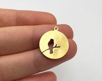 May include: A circular gold-toned metal charm featuring a cutout silhouette of a small bird perched on a branch. The charm has a smooth, matte finish and includes a small circular loop at the top for attachment to jewelry or other accessories.