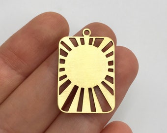 May include: A rectangular gold-toned metal pendant featuring a central solid circle surrounded by radiating cut-out sunburst lines. The piece has a small circular loop at the top for attachment, showcasing a flat, smooth finish with clean edges.