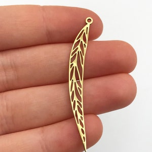 May include: A long, slender, gold-colored metal charm featuring an intricate, openwork leaf or feather pattern. The piece is curved in shape and includes a small circular loop at the top for attachment, showcasing a delicate, filigree-like design.