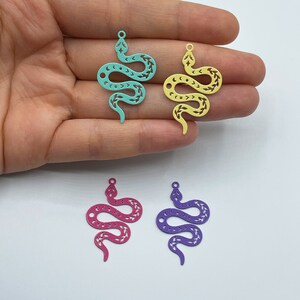 Op de afbeelding: Vier metalen slangenbedels in verschillende kleuren: turquoise, geel, roze en paars. Elke bedel heeft een gedetailleerd slangendesign met een decoratief patroon.