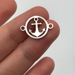 Op de afbeelding: Zilverkleurige metalen bedel met een ankerontwerp in een cirkel. De bedel heeft twee springringen om aan sieraden te bevestigen.