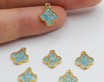 May include: Gold-colored clover-shaped charms with turquoise-colored stones. The charms are small, with a loop at the top for attaching to jewelry. Six charms are visible, with one held in a hand.