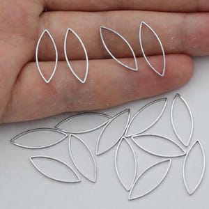 Könnte beinhalten: Eine Sammlung von silberfarbenen, marquiseförmigen offenen Metallrahmen. Diese dünnen, hohlen, augenförmigen Komponenten haben eine glatte, metallische Oberfläche und sind in einer losen Gruppe angeordnet, wobei mehrere Teile auf einer Fläche liegen.