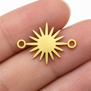 May include: A gold-colored metal sunburst charm featuring a central circular disc with radiating pointed rays and two small circular loops extending horizontally from opposite sides, designed as a connector for jewelry making projects.