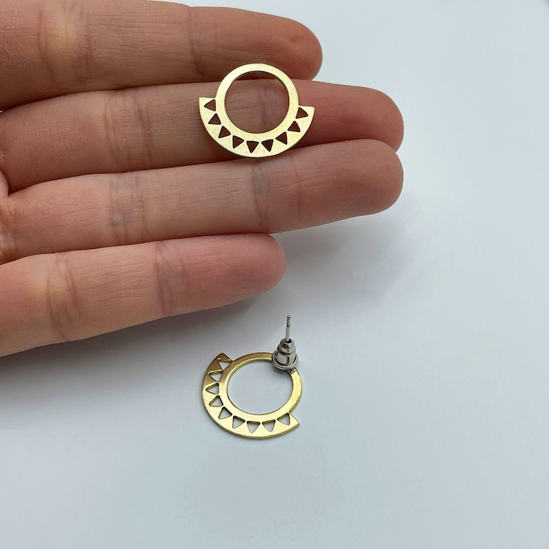 May include: Gold-colored stud earrings. Each earring features a circular design with a semi-circular bottom adorned with triangular cutouts. The earrings have a post and backing for secure wear. The design is geometric and modern.