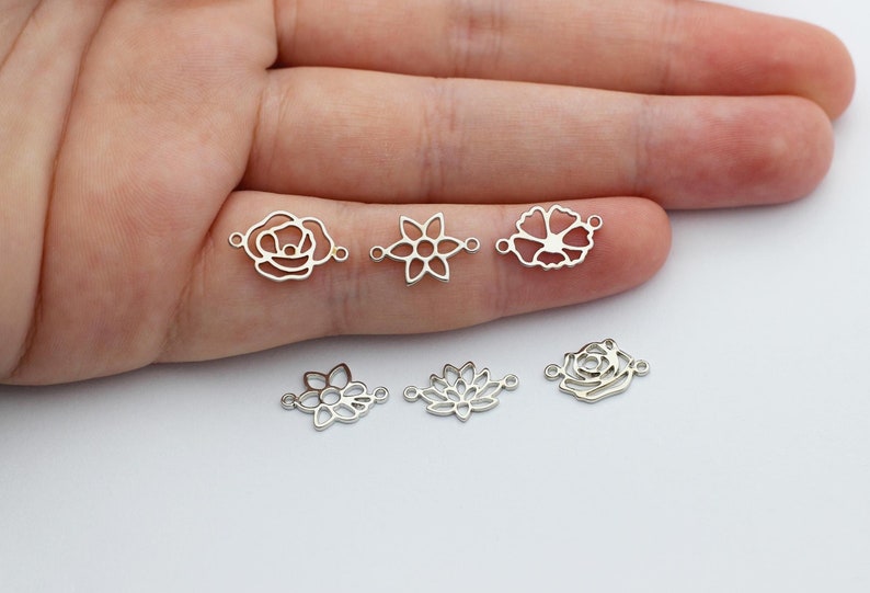 Puede incluir: Seis dijes plateados con forma de flor. Los dijes incluyen dise&ntilde;os de rosa, flor y loto. Cada dije tiene dos peque&ntilde;os bucles para sujetar a joyas u otras manualidades. Los dijes se muestran sobre un fondo claro.