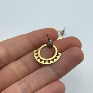 May include: A pair of gold-colored stud earrings. Each earring features a circular design with a semi-circular bottom section adorned with triangular cutouts. The earrings have a post and backing for secure wear.