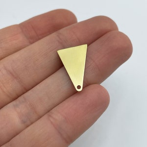 May include: A small, gold-colored triangle charm with a hole at the bottom. The charm is held in a hand against a white background. The charm is made of metal and has a smooth surface.