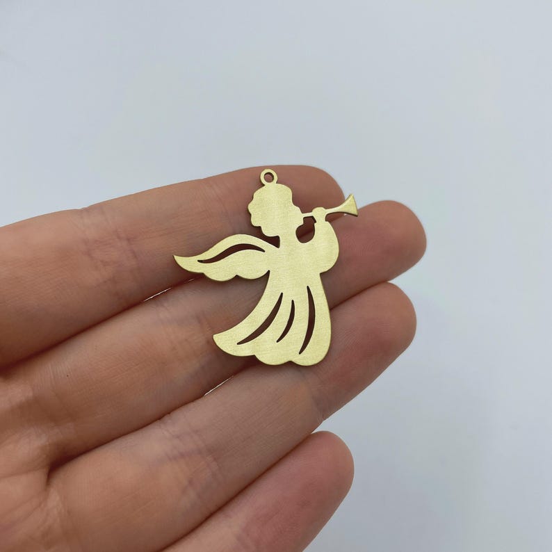 May include: A flat, gold-colored metal charm in the shape of an angel playing a trumpet. The angel figure features a rounded head, outstretched wings, and a flowing gown with cut-out details. A small circular loop is located at the top for hanging.