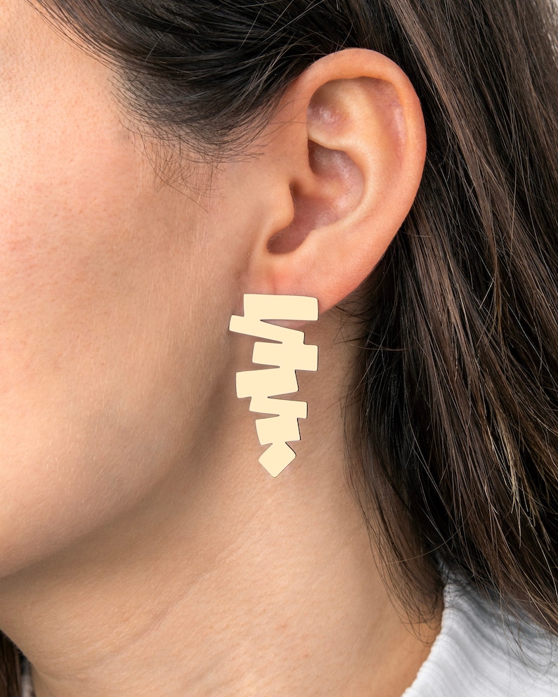 May include: Gold geometric earrings with a stacked, asymmetrical design. The earrings are made of a single piece of metal and have a modern, minimalist style.