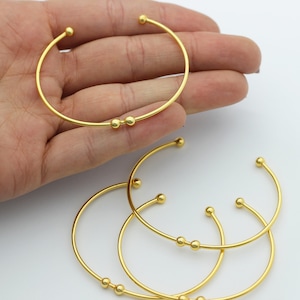 Puede incluir: Cuatro pulseras de brazalete doradas con pequeños acentos redondos. Las pulseras están apiladas una encima de la otra.