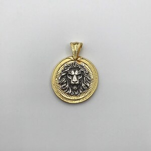 May include: A gold-colored pendant with a detailed lion's head in the center. The lion's head is silver-colored and surrounded by a gold circular frame. The pendant has a small gold loop at the top for a chain.