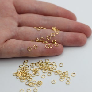 May include: A scattering of small, gold-coloured jump rings on a white surface, with some resting on a person's palm. These circular, shiny components are for jewellery making and are ideal for crafting and design.