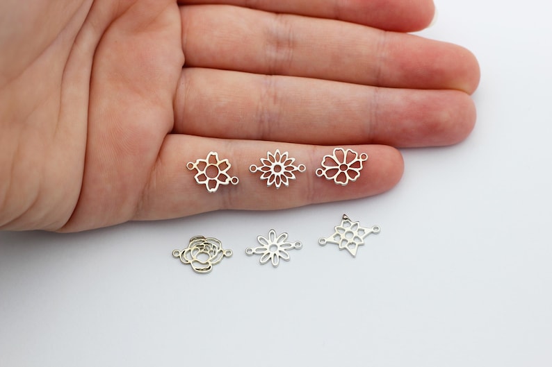 Puede incluir: Seis dijes conectores de metal plateado con varios dise&ntilde;os florales. Los dijes son calados, con formas que incluyen una flor, una margarita y una estrella. Los dijes se muestran sobre una superficie blanca.