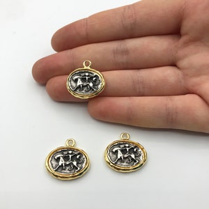 May include: Three gold-toned metal charms featuring a silver-toned design of two figures holding a staff. The charms have a textured, slightly raised edge.