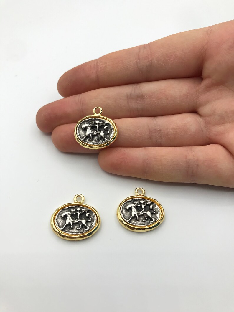 May include: Three gold-toned metal charms featuring a silver-toned design of two figures holding a staff. The charms have a textured, slightly raised edge.