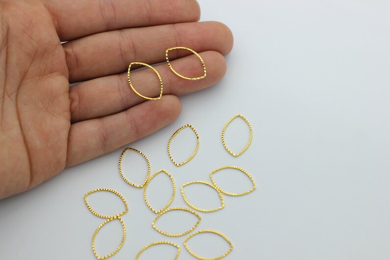 May include: A group of gold-colored metal oval jump rings with a textured surface. The jump rings are scattered on a white surface.