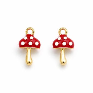 May include: Two mushroom-shaped charms. Each charm features a red cap with white polka dots and a gold-colored stem. The charms have a loop at the top for attaching to jewelry. The charms are set against a white background.