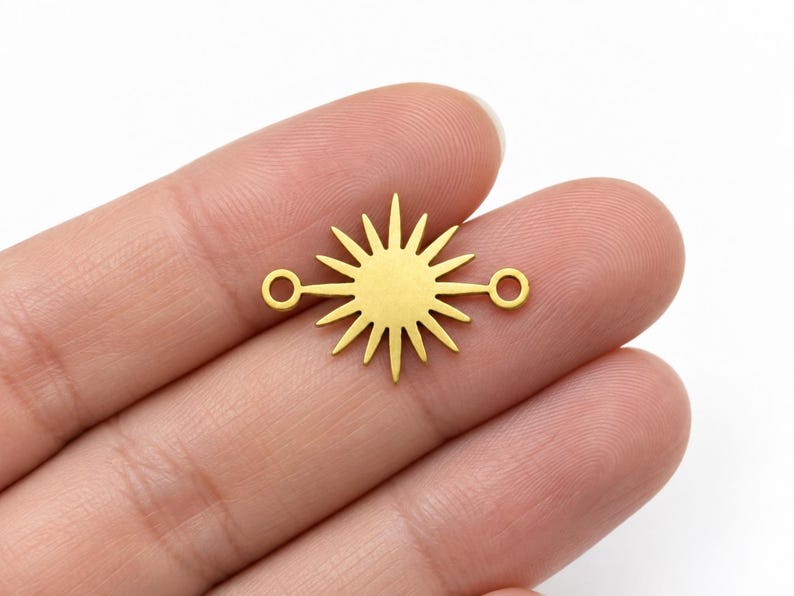 May include: A gold-colored metal sunburst charm featuring a central circular disc with radiating pointed rays and two small circular loops extending horizontally from opposite sides, designed as a connector for jewelry making projects.
