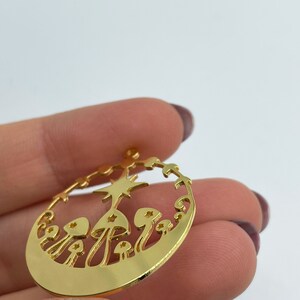May include: Gold-toned metal charm featuring a circular design with a star and mushrooms. The charm has a cut-out design, revealing the background.