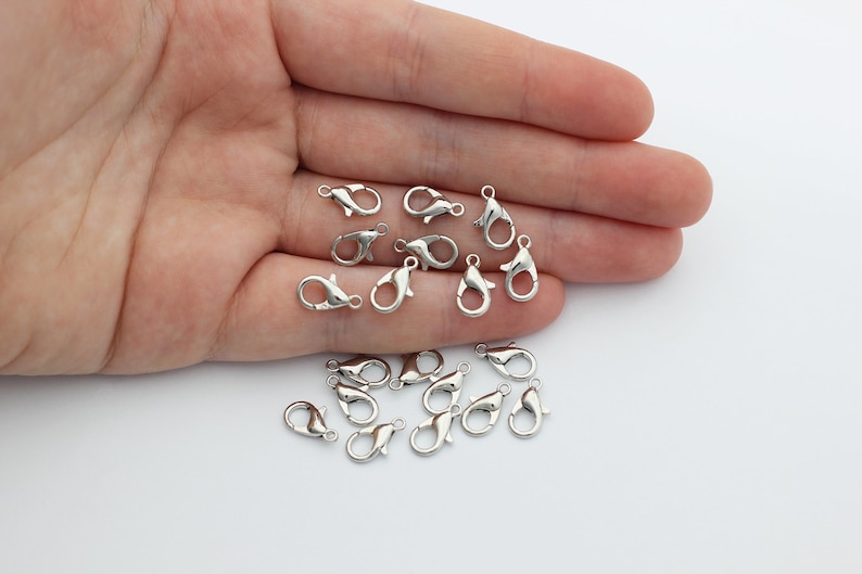 May include: A close-up of a hand holding a pile of silver-toned lobster claw clasps. The clasps are small and delicate, and they are perfect for adding a touch of elegance to jewelry.