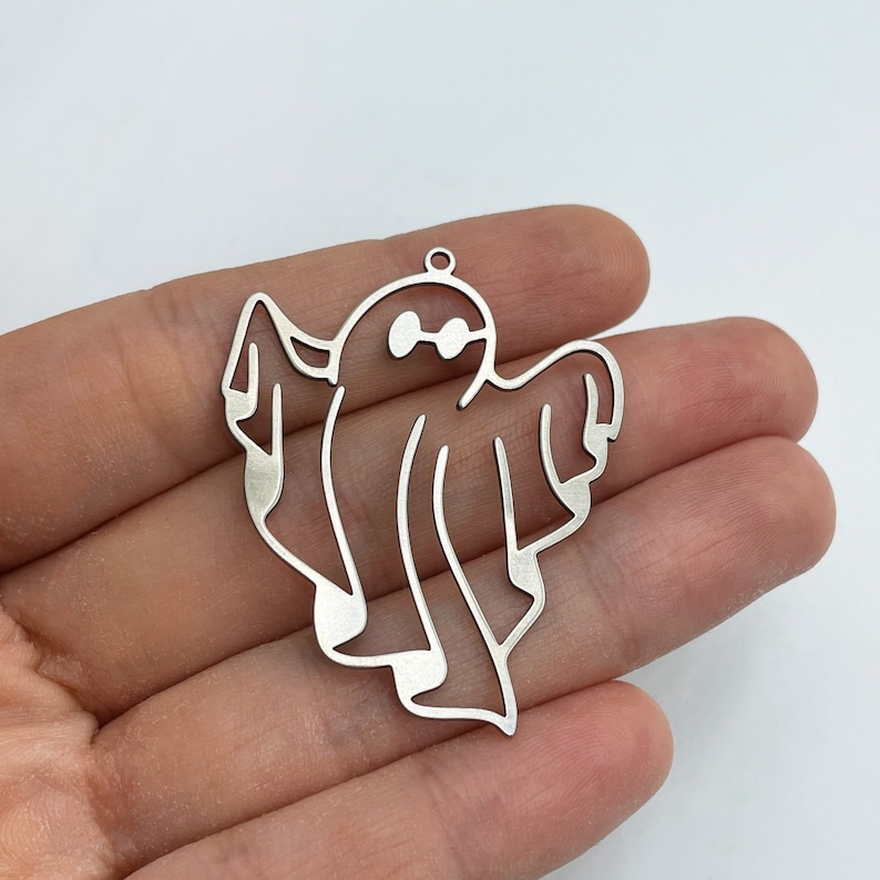 Pode incluir: Um pingente de metal prateado em forma de fantasma com um design de contorno simples. A figura do fantasma apresenta dois recortes circulares para os olhos e um pequeno anel no topo para fixa&ccedil;&atilde;o, com uma superf&iacute;cie lisa e acabamento met&aacute;lico.