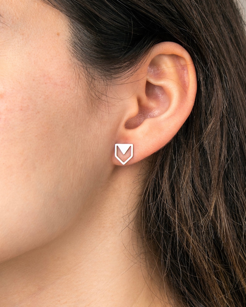 May include: A silver-toned stud earring featuring a geometric design composed of an open-ended pentagonal outline with a downward-pointing triangle centered within the upper portion, securely positioned on the earlobe.