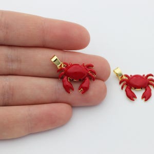 May include: Two red crab pendants with gold-coloured bails. The crab pendants are a bright red colour with detailed claws and body segments. One pendant is held in a hand, and the other is next to it.