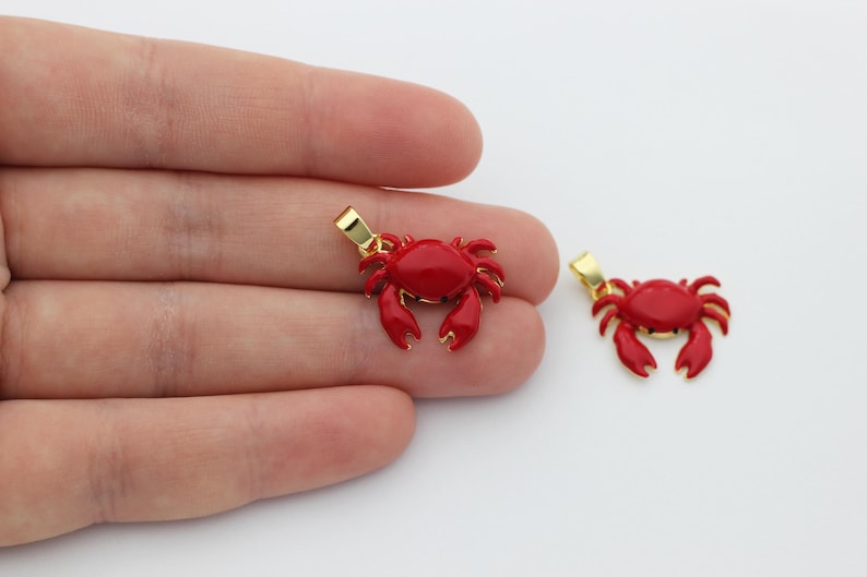 May include: Two red crab pendants with gold-coloured bails. The crab pendants are a bright red colour with detailed claws and body segments. One pendant is held in a hand, and the other is next to it.