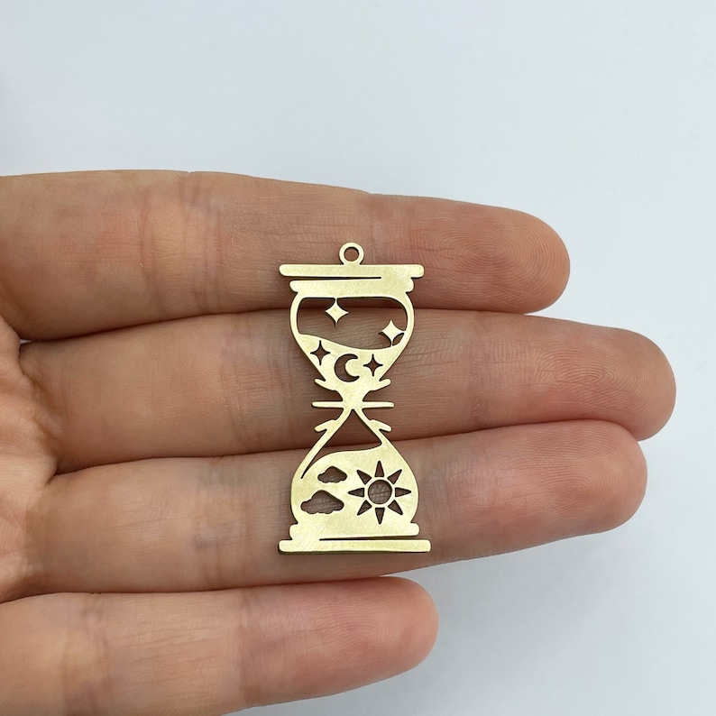 May include: Gold-toned metal hourglass charm with a sun, clouds, moon, and stars design. The hourglass is about 2 inches tall.