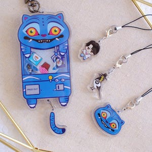 Peut inclure: Ensemble de breloques et porte-clés bleus en acrylique. Le charme principal est une machine à griffes bleue avec un visage de chat. D'autres breloques incluent un visage de chat, un personnage de dessin animé et une figure d'oiseau. Les breloques sont attachées à des cordons noirs.