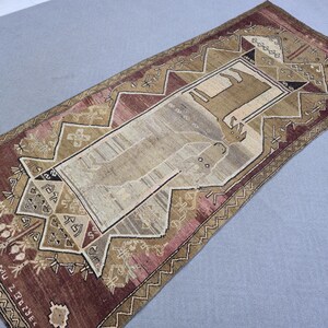 May include: A rectangular, hand-woven rug with a central design featuring a figure in a rectangular frame. The rug has a predominantly brown and beige color palette with accents of pink and cream. Geometric patterns and text border the central design.