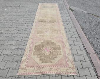Puede incluir: Una alfombra rectangular de estilo vintage, con una combinación de colores beige y rosa. La alfombra presenta patrones geométricos en tonos marrones y rosas, con un borde rosa. Es larga y estrecha, ideal para pasillos o entradas, unos 200 cm de largo.