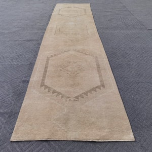 May include: A long, narrow runner rug in a light beige color featuring three repeating hexagonal geometric patterns centered along its length. The design includes subtle, darker brown linear motifs within each hexagon and a serrated border.