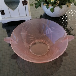 Op de afbeelding: Een roze glazen kom met twee handvatten en een swirl-ontwerp. De kom is leeg en staat op een zwart oppervlak.