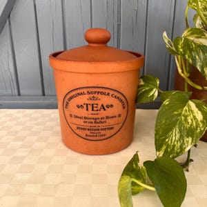 May include: A terracotta canister with a matching lid, featuring black printed text that reads The Original Suffolk Canister, TEA, Ideal Storage at Home or on Safari, Made by Henry Watson Pottery, England, Founded 1800.
