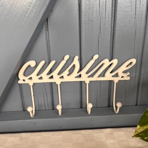 May include: Off-white metal wall hook with the word "cuisine" in cursive script. The hook has four curved hooks below the word. The background is a light blue wooden surface.