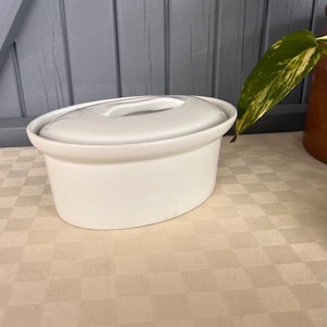 May include: A white ceramic oval casserole dish with a matching lid. The lid has a handle. The dish is on a beige checkered tablecloth. A green and yellow leaf is visible in the background.