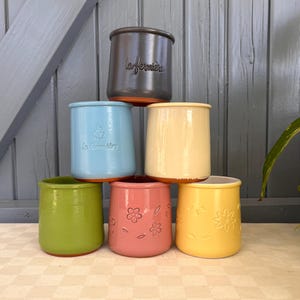Può includere: Una pila di sei vasetti in ceramica in vari colori: grigio scuro, azzurro, crema, verde, rosa e giallo. Il vasetto grigio scuro ha il testo "la fermiere". Il vasetto rosa ha un motivo floreale. I vasetti sono impilati su una superficie a scacchi.