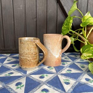 Może przedstawiać: Dwa ręcznie robione ceramiczne kubki z brązową i białą, kropkowaną glazurą. Kubki mają zaokrąglony kształt i uchwyt. Stoją na niebiesko-białym wzorzystym obrusie.
