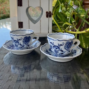 Op de afbeelding: Twee witte kopjes en schotels met een blauw bloemmotief. De kopjes hebben geschulpte randen en kleine oren. De schotels zijn rond met een bijpassend blauw bloemmotief. De set staat op een reflecterend oppervlak.