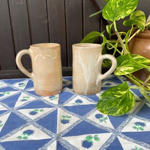 Może przedstawiać: Dwa ceramiczne kubki z białą glazurą i brązowym, kropkowanym wykończeniem. Kubki mają uchwyt i stoją na niebiesko-białym wzorzystym obrusie.